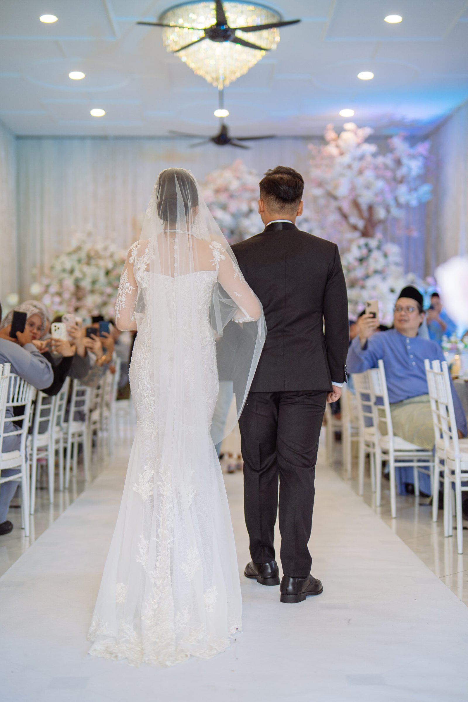 ack view of a bride in a white lace gown and groom in a black suit walking down the aisle at a modern Malay wedding at The Agam.