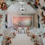 Romantic wedding aisle adorned with floral arrangements leading to an intimate ceremonial setup at The Agam, framed by an arch of blush and soft pink flowers.