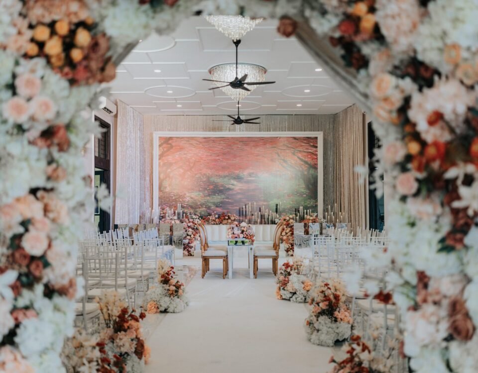 Romantic wedding aisle adorned with floral arrangements leading to an intimate ceremonial setup at The Agam, framed by an arch of blush and soft pink flowers.